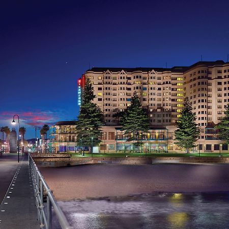 Stamford Grand Adelaide Hotel Glenelg Eksteriør billede The photo shows a waterfront scene at dusk, featuring a large building lit up against a darkening sky. The architecture appears modern and prominent, with several windows illuminated. In the foreground, there is a walkway or pier with fencing along t