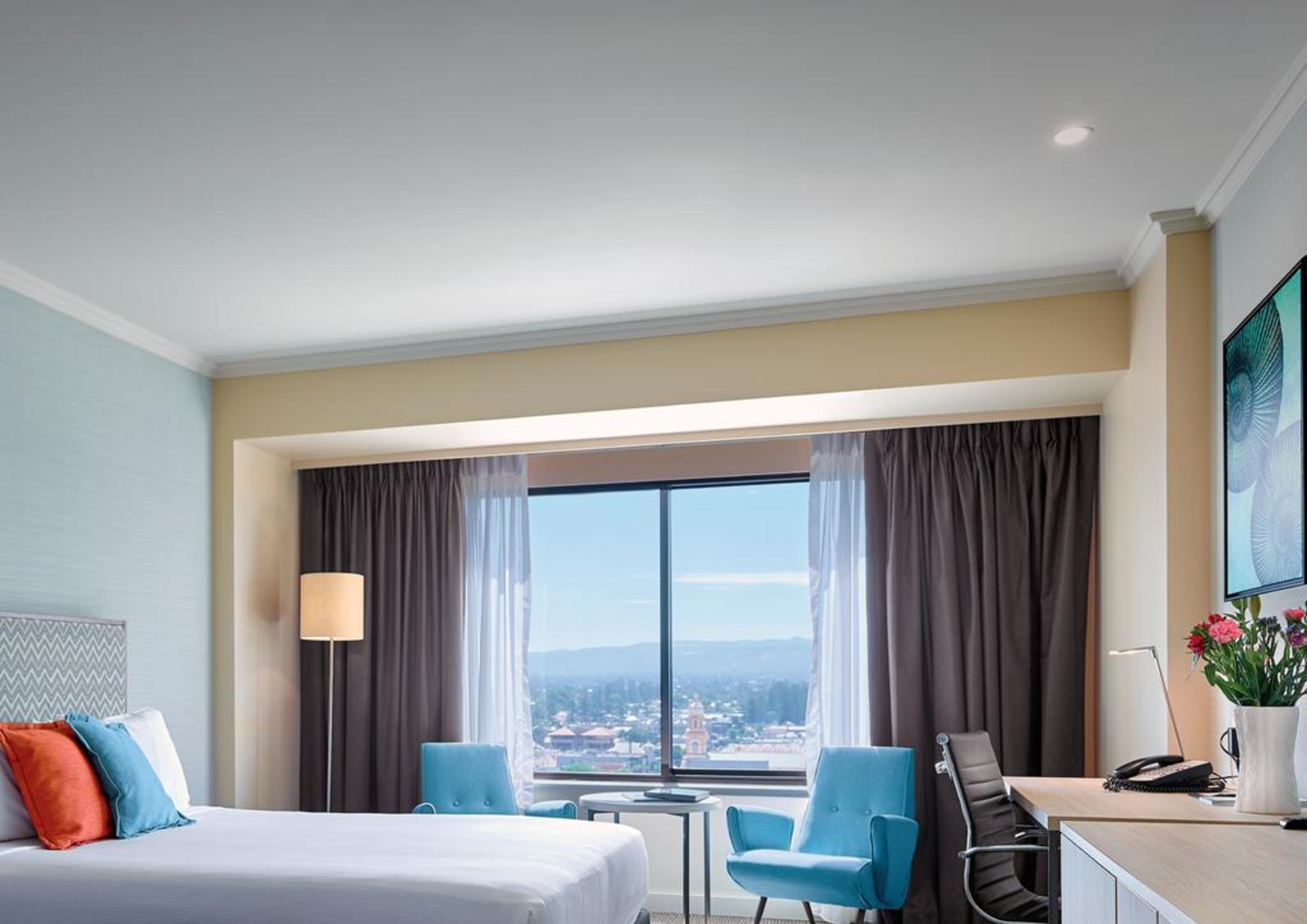 Stamford Grand Adelaide Hotel Glenelg Eksteriør billede The photo shows a modern hotel room featuring a queen-sized bed with white linens and colorful decorative pillows. There are two blue chairs beside a small table positioned near a large window that provides a view of the outside, with sheer and dark 