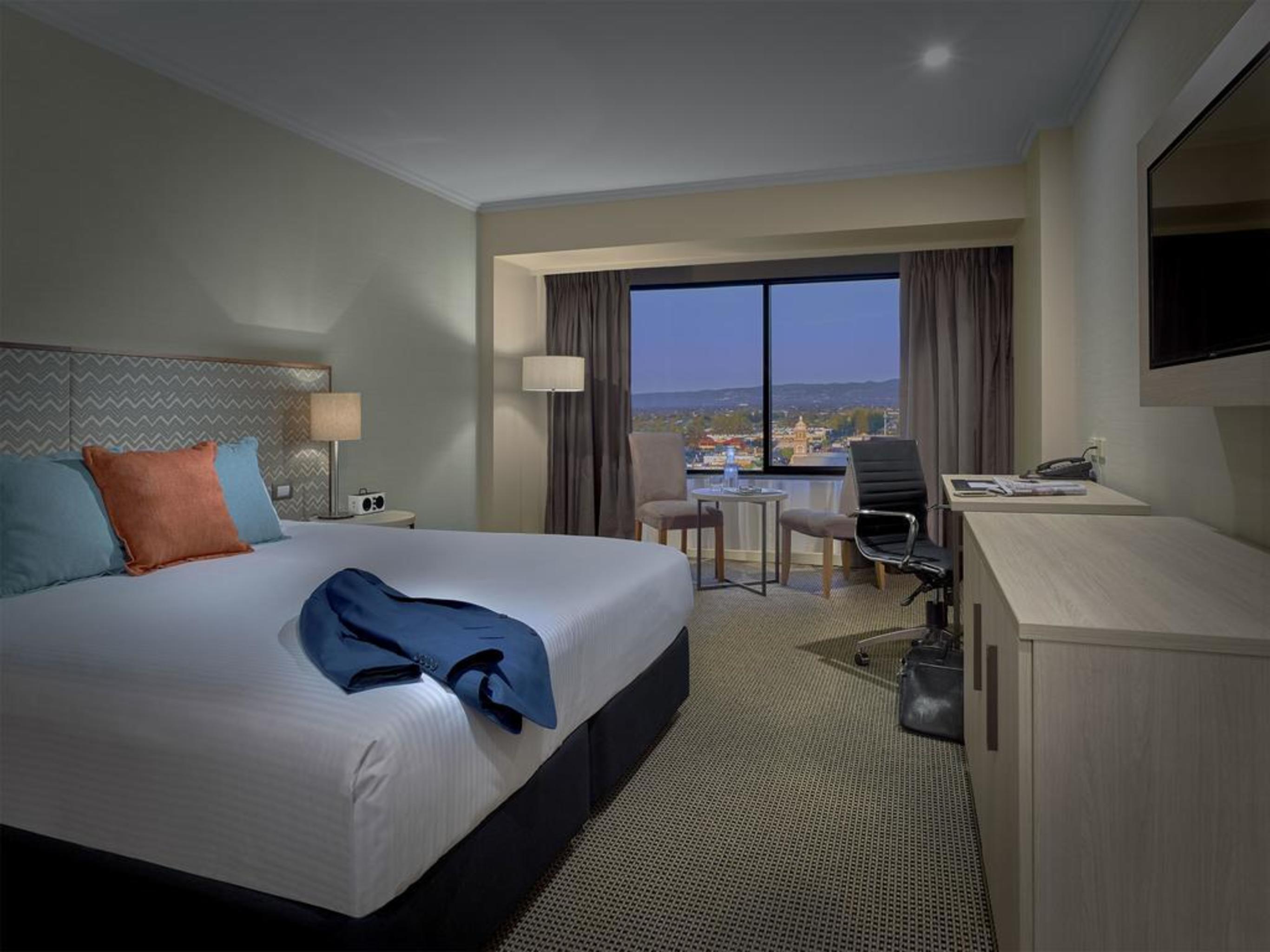Stamford Grand Adelaide Hotel Glenelg Eksteriør billede The photo shows a modern hotel room. It features a large double bed with white bedding and decorative pillows in orange and blue. There's a nightstand with lamps on either side of the bed. A large window overlooks a view outside, and there's a desk a
