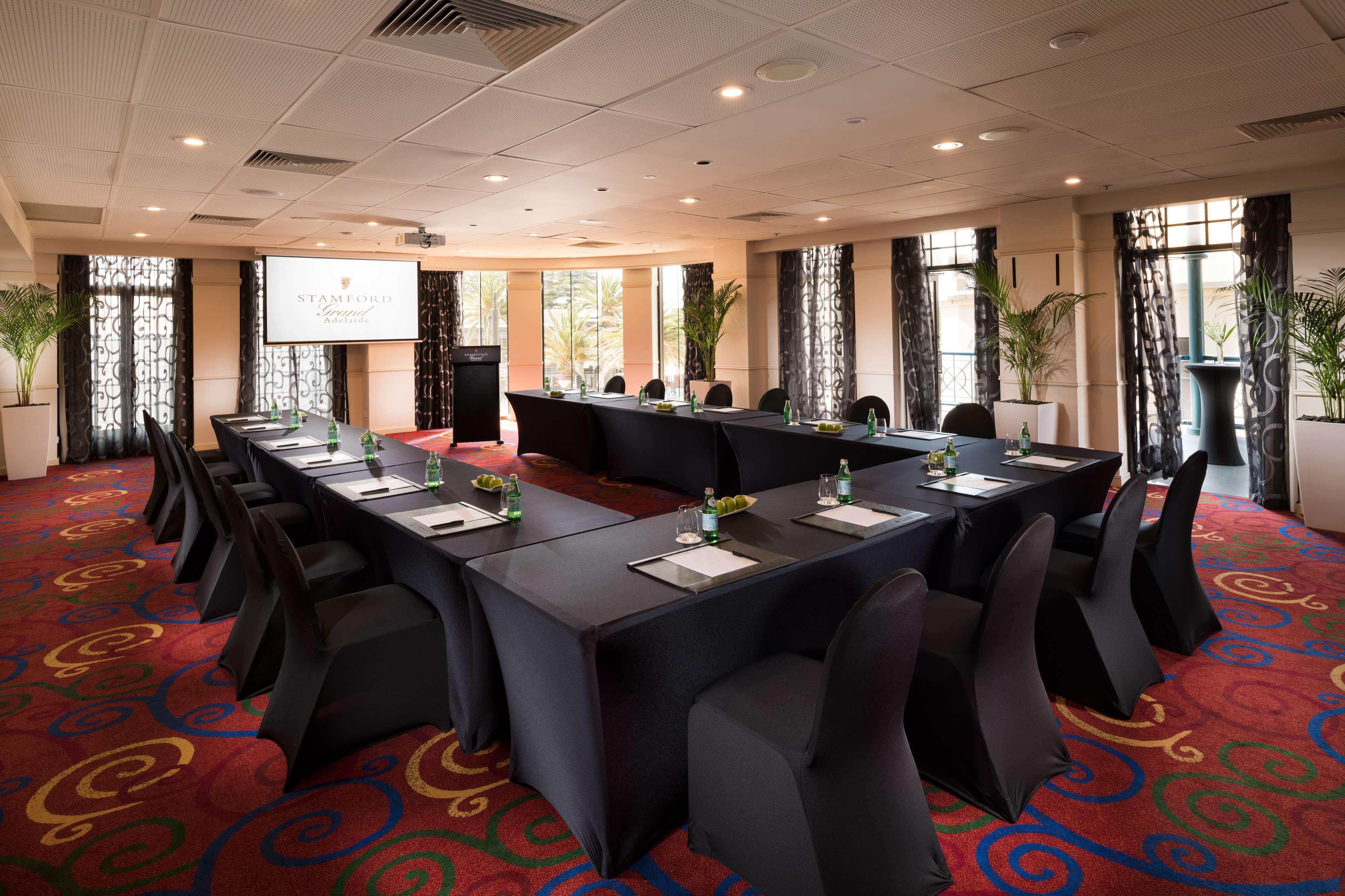 Stamford Grand Adelaide Hotel Glenelg Eksteriør billede The photo shows a conference room set up for a meeting or presentation. There are several tables arranged in a U-shape, covered with black tablecloths. Each table has a notepad and a small bottle of water on it. The chairs around the tables are also 
