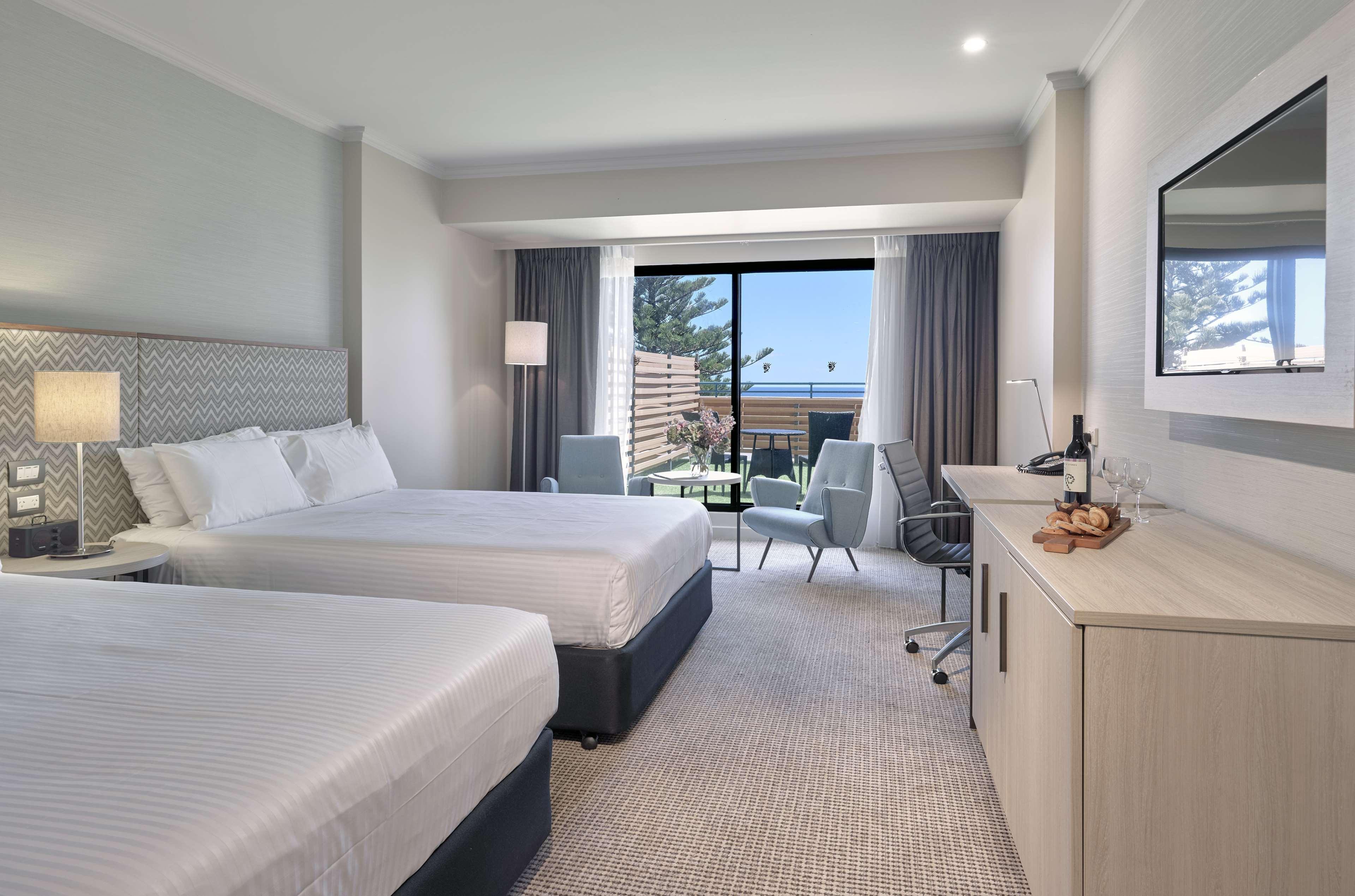 Stamford Grand Adelaide Hotel Glenelg Eksteriør billede The photo shows a modern hotel room featuring two beds with white linens. There's a large window that provides natural light and a view of a balcony area. The room includes two chairs and a small table, creating a cozy sitting area. A television is m