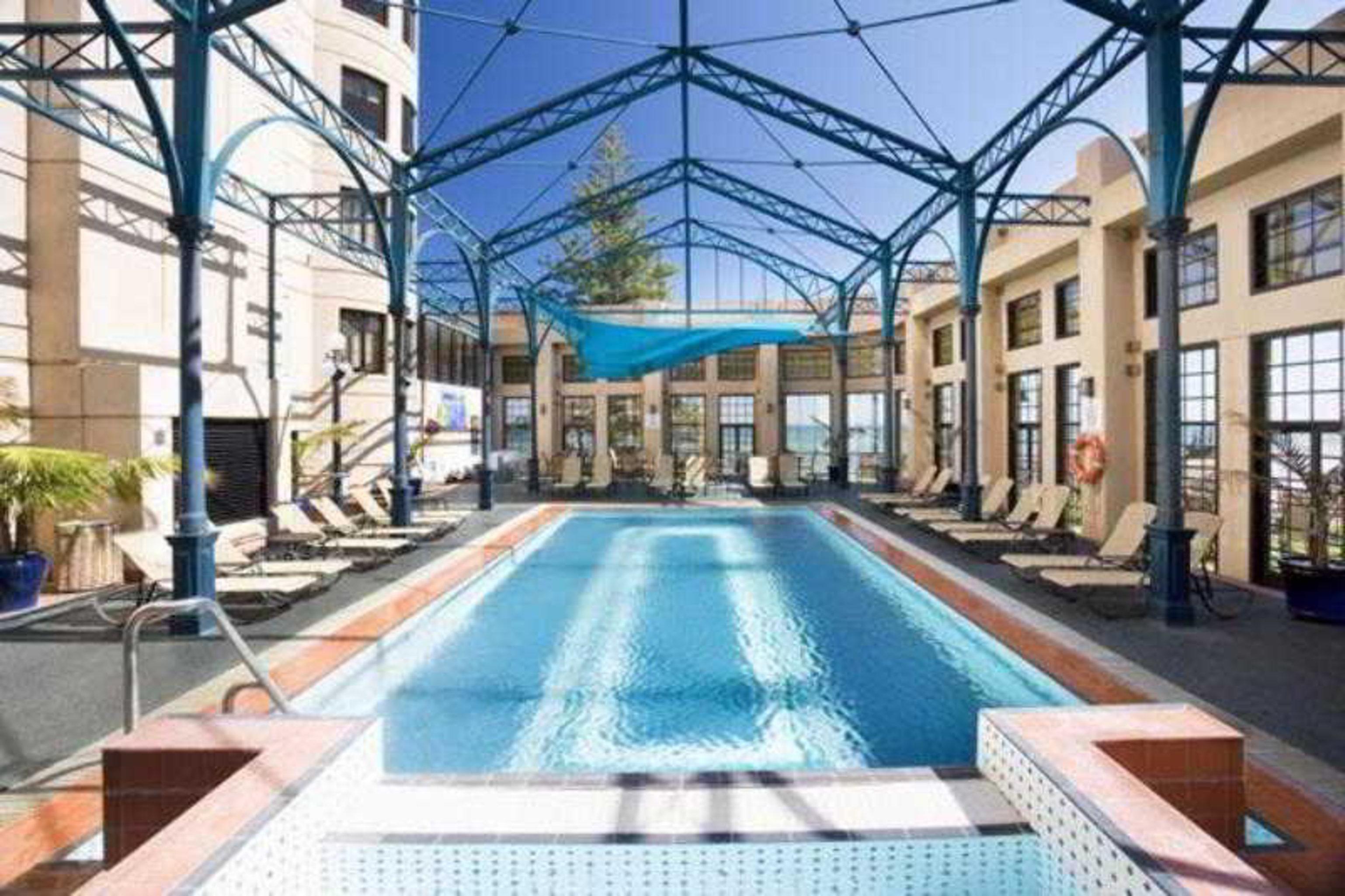 Stamford Grand Adelaide Hotel Glenelg Faciliteter billede The photo shows an indoor swimming pool area. The pool is rectangular and features a blue cover above, supported by a metal frame. Surrounding the pool are several lounge chairs, and the space is well-lit with large windows allowing natural light to 